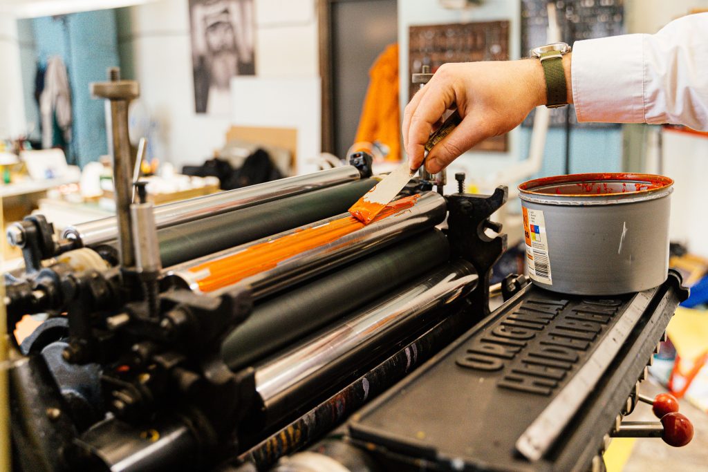 Document Printing A detailed view of a hand applying orange ink to a vintage printing press roller indoors.